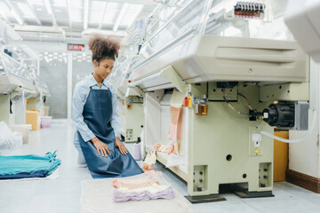 curly hair american female designer Looking at the fabric she has designed near the sewing machine in a large weaving industry There are many machines working. She's wearing a uniform.