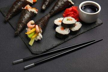 Slate board with tasty sushi cones, rolls, soy sauce, ginger and chopsticks on dark background, closeup