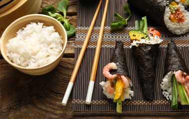 Bamboo mat with tasty sushi cones, chopsticks and boiled rice on wooden background, closeup