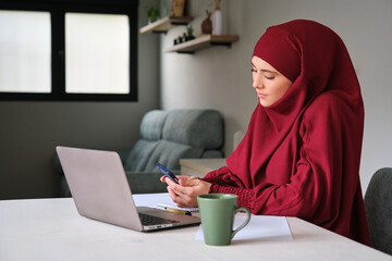 Muslim young woman in hijab using phone while working at home.