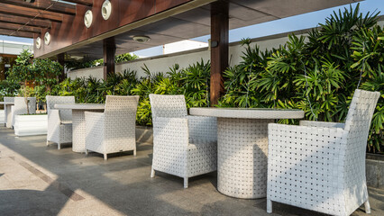 White wicker chairs and tables stand in a row under a canopy in a cafe on an outdoor terrace. Tile floor. Green vegetation nearby. - obrazy, fototapety, plakaty