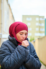 Muslim young woman in hijab and coat coughing in street.