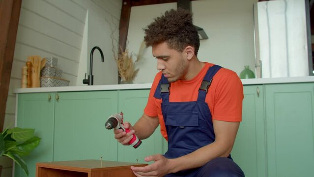 Puzzled attractive African American man in protective workwear having problems using power tool , expressing frustration and confusion while assembling DIY furniture with electric cordless screwdriver