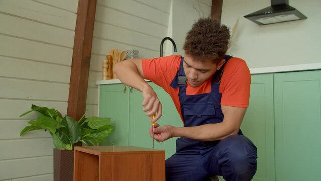 Portrait of skillful handsome black handyman in protective workwear doing DIY job, repairing old furniture, tightening self tapping screws with screwdriver in domestic room.