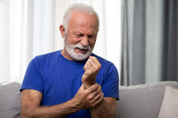 Elderly man holding his wrist, suffering from arthritis pain in hand, sitting on sofa at home - obrazy, fototapety, plakaty