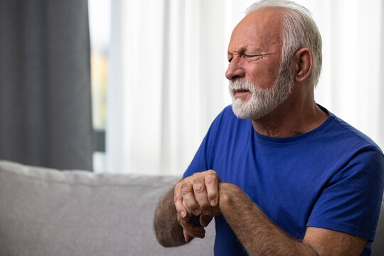 Elderly Man Holding His Wrist, Suffering From Arthritis Pain In Hand, Sitting On Sofa At Home