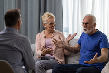 Unhappy senior couple arguing, having fight, disagreement at psychologists office. Frustrated elderly wife and husband discussing relationship problems with their therapist - 602222521