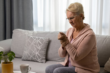 Elderly woman holding her wrist, suffering from arthritis pain in hand, sitting on sofa at home