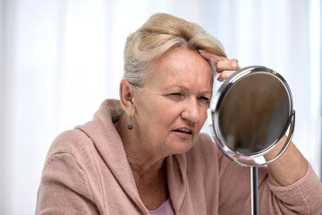 Anxious senior woman looking in mirror and feeling worried about face wrinkles, skin elasticity or hair loss. Pensive female thinking of facial injection, cosmetic procedures or plastic surgery - 602222714