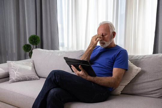 Senior man using digital tablet computer at home and having eyestrain or suffering from astigmatism and bad vision. Eyesight problems concept.