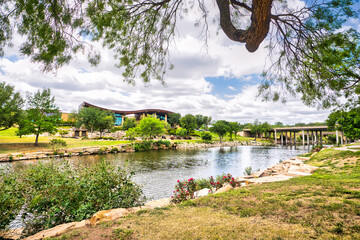 A visitor center on the other side of a tranquil river in San Angelo, Texas, surrounded by lush grass and trees with wispy clouds dotting a bright blue sky.