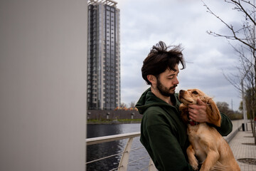 A bearded man standing near the river holds a cocker spaniel in his arms and scratches his neck. A man in a green jacket hugs his dog during a morning walk in the park.