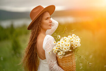 A middle-aged woman in a white dress and brown hat stands on a green field and holds a basket in her hands with a large bouquet of daisies. In the background there are mountains and a lake.