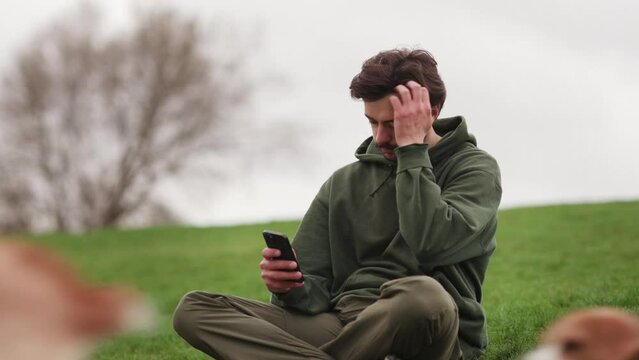 A man sits on the grass and browses social networks on his smartphone. A brunette man with a phone in his hands sits on the grass while dogs run around him.