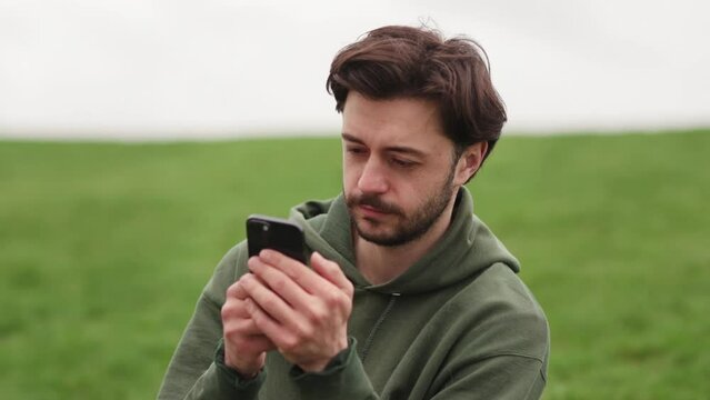 The cocker spaniel runs up to his owner, who is sitting on the grass in the park and looking at something on the phone. A man sits on the grass and browses social networks, while his dog runs near him