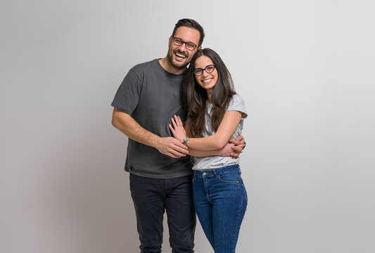 Portrait of loving young couple embracing and holding hands while posing ecstatically against background. Charming girlfriend and boyfriend wearing eyeglasses standing together and looking at camera