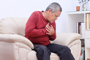 Elderly Asian male clutching his left chest from acute pain while sitting at the couch in the living room. 