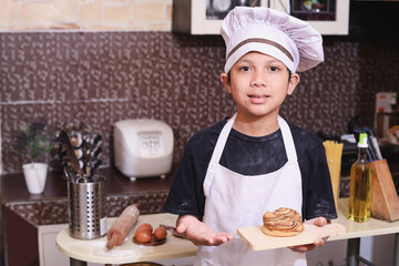 Young little and cute Asian kid in chef uniform presenting and showing tray of pastry smiling happy and proud in learning baking concept at modern home kitchen