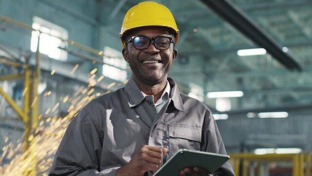 African American architecor in protective casque hold tablet. Male engineer is posing and smiling at camera. Worker at background of construction sity. Variety of industrial professions.