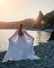 Fototapeta na wymiar Mysterious young woman with braids in long white dress alone on the beach. Sunset over the sea with rocky volcanic cliff. Abstract nature summer ocean sea background.