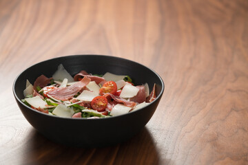 Salad with prosciutto, tomato, arugula leaves and parmesan cheese in black bowl on walnut