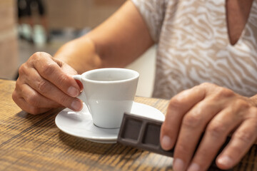 Old caucasian senior woman holding dark chocolate bar in hand while enjoying cup of espresso coffee - break time with hot aromatic drink