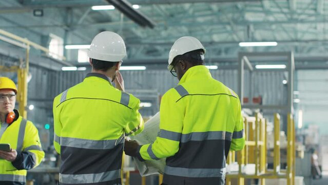 Engineers are going at construction site. Workers are shooted from back and communicate together. Manufacture employee dressed up in reflective vest. African American male holds blueprint.