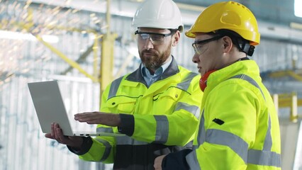 Two employees at construction site. Engineer is holding laptop and explain tasks to contractor. Building workers create plan. Two men at backgrounf of equipment and welding sparks flying.