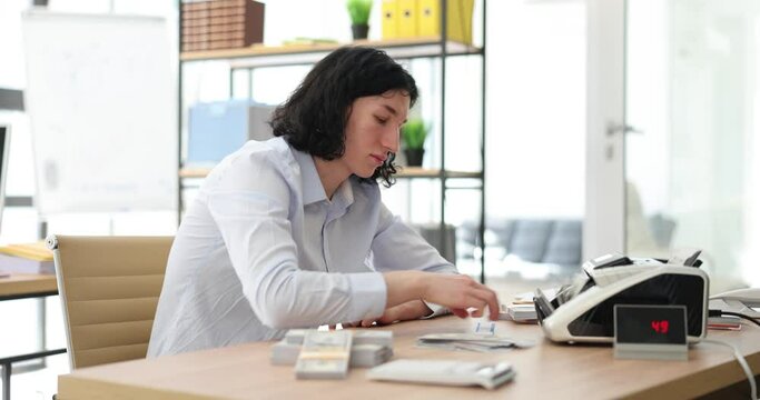 Male employee puts stack of dollar banknotes into bill counter. Long-haired man counts cash and puts packed wad of money on wooden table in office