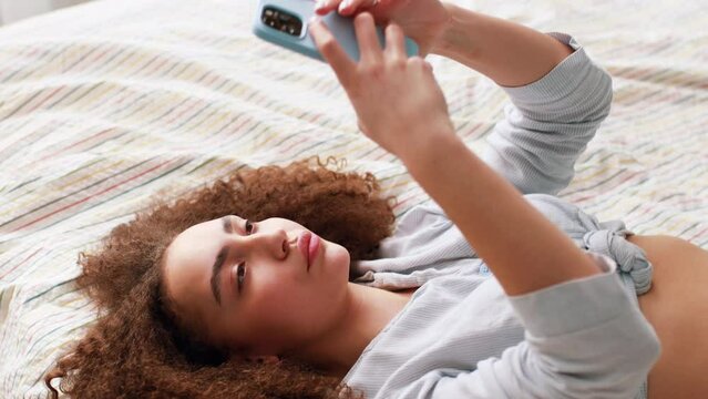 Young beautiful woman with brown wavy, curly hair using her smart phone and lying in bed in the morning. Girl takes morning selfie on smartphone. Top view, slow motion lifestyle free spare time