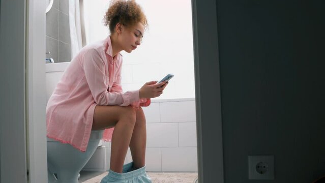 Young woman using her cell phone while sitting on toilet, texting message, sms, touching screen of ceell phone chatting conversation slow motion