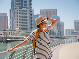 Attractive traveler asian woman with yellow backgpack walking on promenade in Dubai Marina district. Travel destinations and tourist lifestyle in UAE