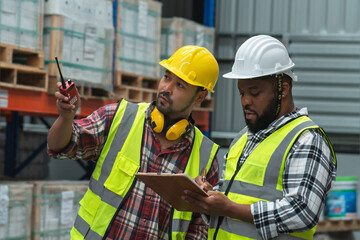 Serious Asian foreman holding walkie talkie and African engineer in safety hat look at inventory, working at site, listing problem on clipboard to discuss, inspect plan for new building at warehouse