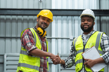 Happy Asian foreman and African engineer in safety hat shaking hand and smiling at camera, working at site, holding clipboard to list problem, good cooperation of colleagues at warehouse