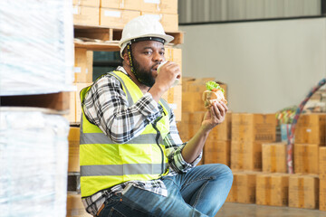 Happy African middle aged engineer worker beard man wear safety hat, eating burger, coffee in lunch break at warehouse. Foreman relaxing having lunch at workplace