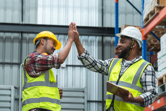 Asian foreman and African engineer in safety hat giving high five and smiling at each other, working at site, listing problem on clipboard to discuss, inspect plan for build a building at warehouse - Powered by Adobe
