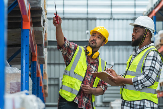 Serious Asian foreman holding walkie talkie and African engineer in safety hat look at inventory, working at site, listing problem on clipboard to discuss, inspect plan for new building at warehouse