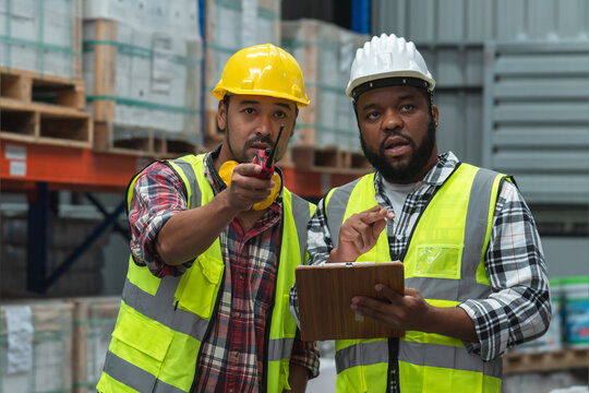Serious Asian foreman holding walkie talkie and African engineer in safety hat look at inventory, working at site, listing problem on clipboard to discuss, inspect plan for new building at warehouse