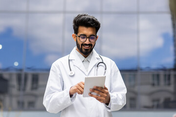 Young successful Hindu student in white medical coat walks outside clinic, doctor intern trainee with tablet computer in hands, browsing online educational material. - Powered by Adobe