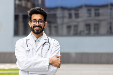 Portrait of a happy and successful Hindu doctor, a young medical practitioner smiling and looking at the camera outside a medical clinic, a man in a medical gown with crossed arms and a stethoscope.