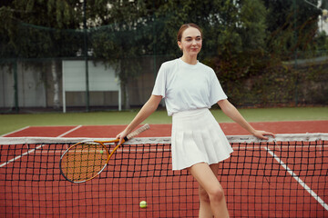 Fashionable woman in white clothing with tennis racket posing at tennis net on court. Sports Fashion