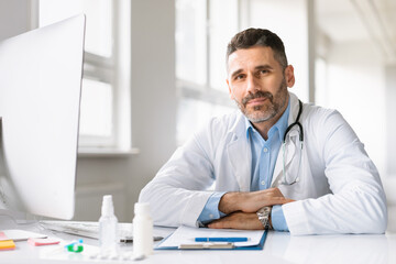 Portrait of professional male doctor in white workwear sitting at workplace, therapist wearing coat and stethoscope