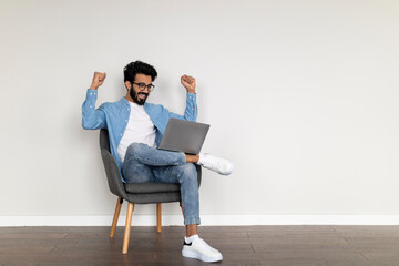 Joyful Indian Male Using Laptop And Shaking Fists While Sitting In Chair