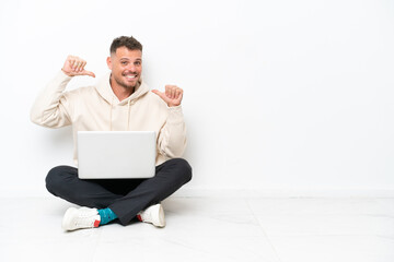 Young caucasian man with a laptop sitting on the floor isolated on white background proud and self-satisfied