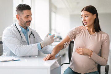 Young pregnant woman scared of vaccine injection, looking at syringe with fear at hospital