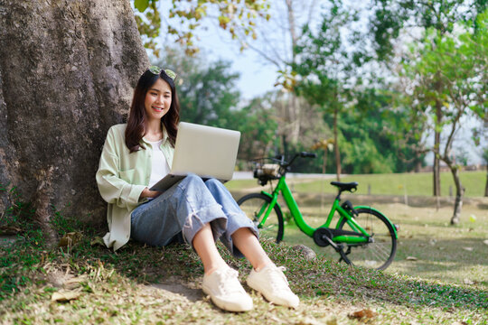 Women sitting under tree to working and surfing social media on laptop after cycling in the park
