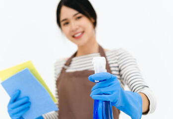 Housekeeper woman holding blue bottle with cleaner liquid and sponge in hands, female wearing white t shirt ,brow apron and blue gloves, looking smiling directly at camera, posing against white wall.