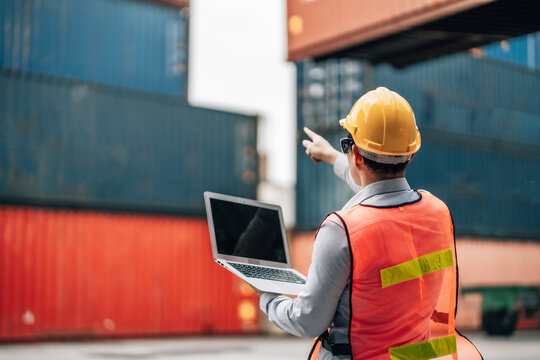 Asian technician dock worker in protective safety jumpsuit uniform and with hardhat and use laptop computer at cargo container shipping warehouse. transportation import,export logistic industrial