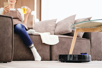 Black robotic vacuum cleaner cleaning the floor while woman sitting on sofa and relax.Young woman...