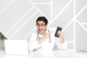 Young handsome physician in white medical gown holding phone and working on laptop in his modern...
